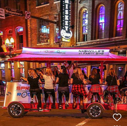 Downtown Nashville nightlife: red party bike with seated riders pedaling at a bar-style counter beneath neon-lit brick buildings