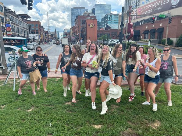 Cheerful bachelorette group in denim shorts and cowboy boots laughing on a grassy patch beside a busy downtown entertainment street lined with brick bars, signs, and modern high-rises under a partly cloudy sky.