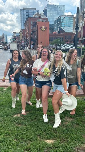Cheerful bachelorette group in denim shorts and cowboy boots laughing on a grassy patch beside a busy downtown entertainment street lined with brick bars, signs, and modern high-rises under a partly cloudy sky.