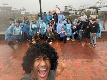 Cheerful selfie of a large group in blue rain ponchos on a rainy riverfront promenade, with a stadium and bridge in the background.