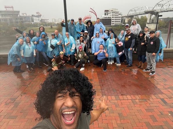 Cheerful selfie of a large group in blue rain ponchos on a rainy riverfront promenade, with a stadium and bridge in the background.