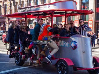 Group of friends laughing and waving while pedaling a colorful party bike down a busy downtown Nashville street with brick storefronts.