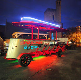Neon-lit red pedal party bike glowing under a canopy at night in Nashville, parked by a stone church tower — festive nightlife pedal-pub scene.