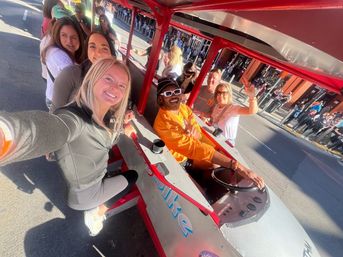Smiling group selfie aboard a red pedal-powered party bike on a sunny downtown street, passengers waving while crowds line the sidewalk.