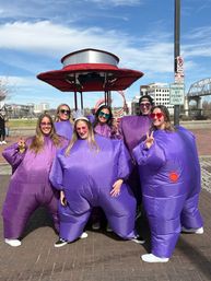 Six people in inflatable purple costumes and colorful sunglasses, one wearing a white tiara, smiling and posing on a sunny urban riverfront promenade with a red canopy, pedestrian bridge and city buildings in the background.