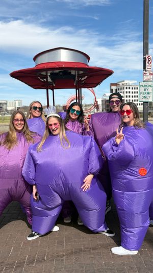 Six people in inflatable purple costumes and colorful sunglasses, one wearing a white tiara, smiling and posing on a sunny urban riverfront promenade with a red canopy, pedestrian bridge and city buildings in the background.