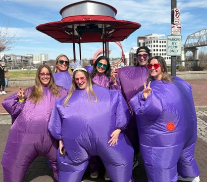 Six people in inflatable purple costumes and colorful sunglasses, one wearing a white tiara, smiling and posing on a sunny urban riverfront promenade with a red canopy, pedestrian bridge and city buildings in the background.