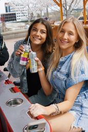 Two smiling women in denim outfits toast canned sparkling drinks on a sunny riverside patio with blossoming trees and city buildings in the background.