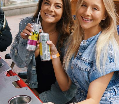 Two smiling women in denim outfits toast canned sparkling drinks on a sunny riverside patio with blossoming trees and city buildings in the background.