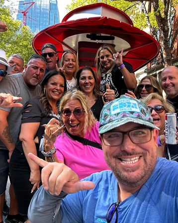 Smiling group selfie of adults on a red‑canopied party bike (pedal pub) in a sunny downtown street with trees and a glass skyscraper in the background