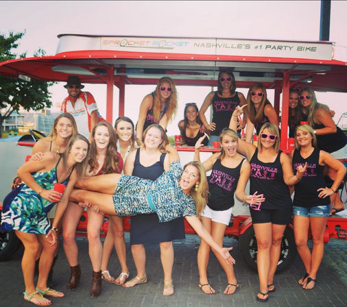 Bachelorette group of women posing on a red Nashville party bike, wearing matching tank tops and sunglasses with one friend lifted playfully