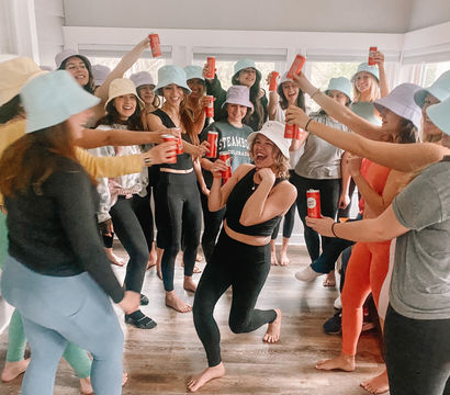 Group of people in colorful bucket hats and activewear cheering indoors, raising red cans toward a smiling person striking a playful pose in the center on a hardwood floor.