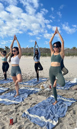 Group of women practicing tree pose in a beach yoga class on a sandy shore, balancing on blue striped towels under a bright blue sky with fluffy clouds