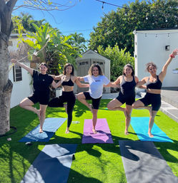 Five women holding tree pose on colorful yoga mats in a sunny tropical backyard, arms linked and smiling during an outdoor group yoga session.