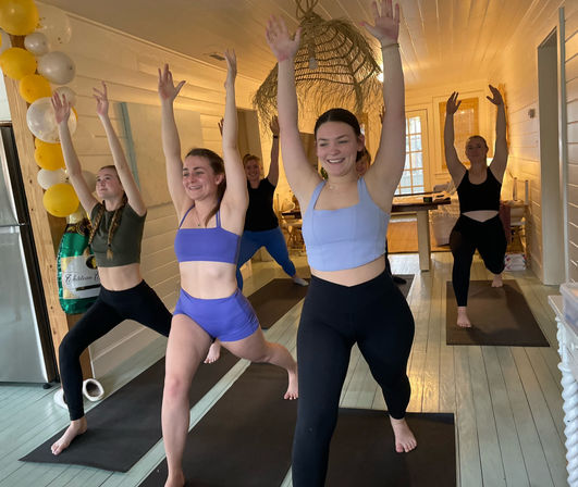 Group of women practicing yoga warrior lunge in a bright wooden studio, smiling with arms raised on black mats, yellow-and-white balloon garland in the corner.