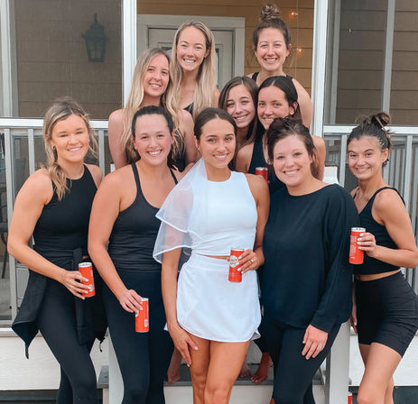 Smiling group of women on a house porch celebrating a bachelorette — bride in a white mini dress and veil holding a red can, friends in black activewear gathered around her.