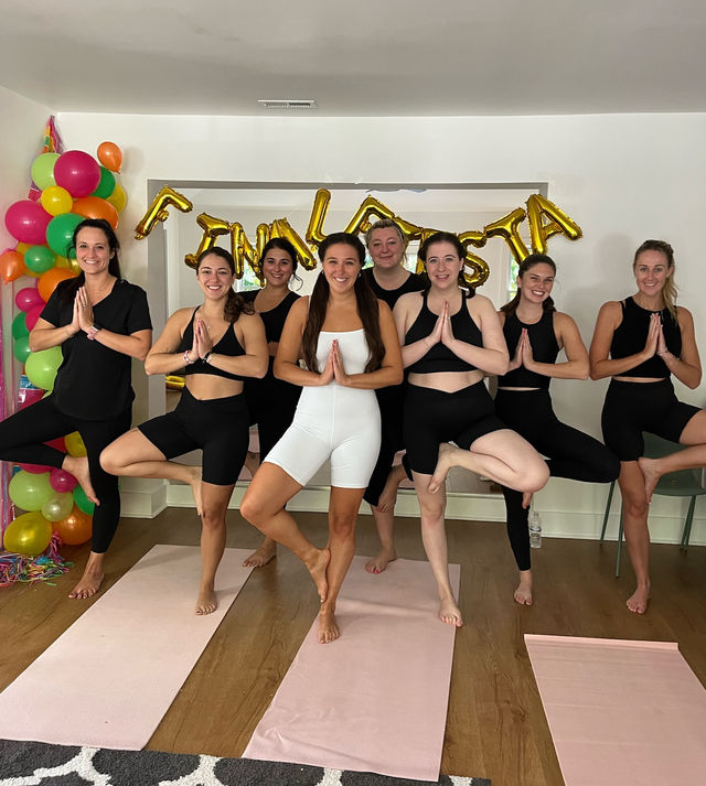Eight women in activewear balancing in tree pose on pink mats in a bright indoor yoga space with colorful balloons and gold letter celebration balloons behind them