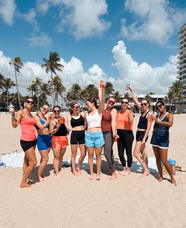 Group of women in activewear raising canned drinks on a sunny palm-lined beach with blue sky and fluffy clouds