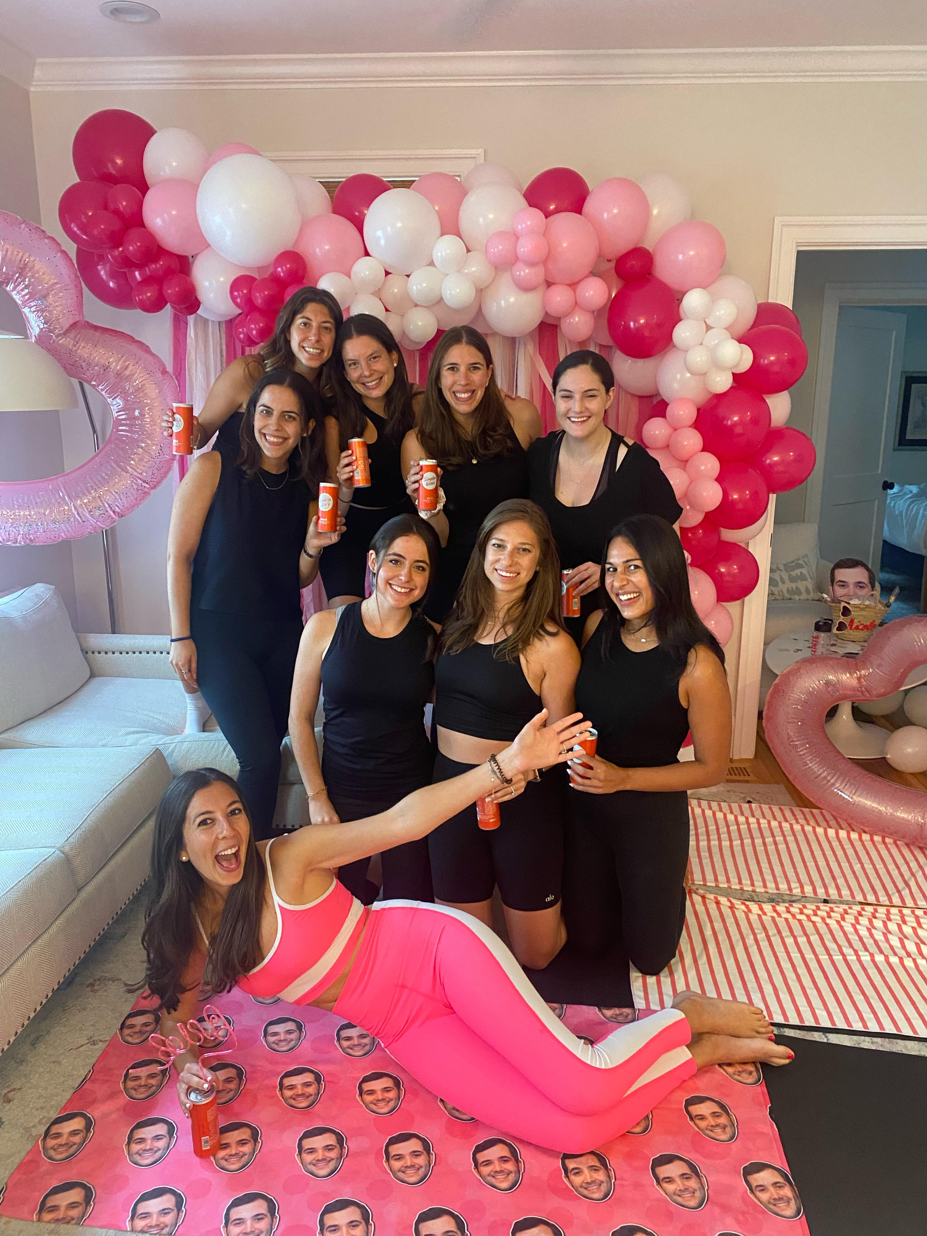 Smiling group of nine women in a living room, most wearing black activewear and one in bright pink workout set, posing in front of a pink-and-white balloon arch with heart-shaped inflatables, holding orange drink cans and lying on a mat printed with repeating face images.