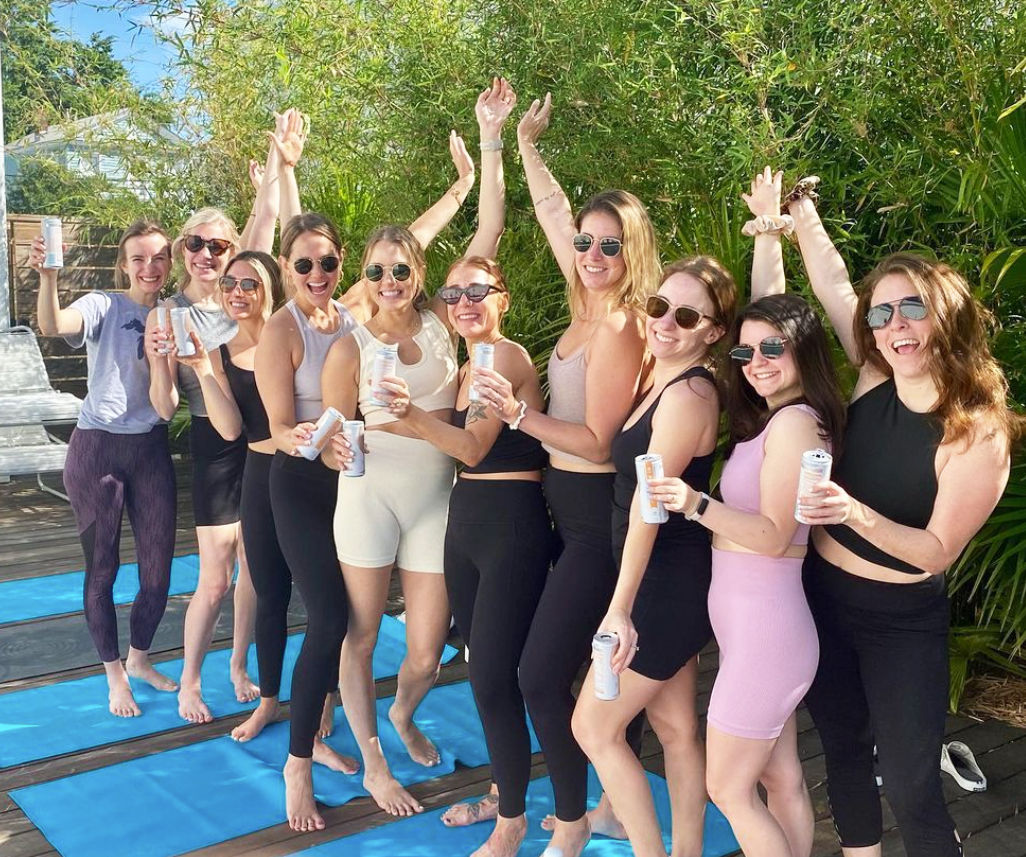 Smiling group of women in activewear on blue yoga mats on a sunny backyard deck, holding canned drinks and raising their arms in front of bamboo greenery