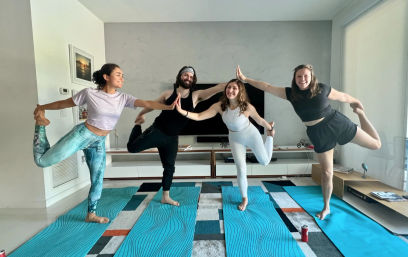 Four people practicing home yoga in a bright modern living room — balancing in dancer pose on blue mats, touching hands in the center and smiling.