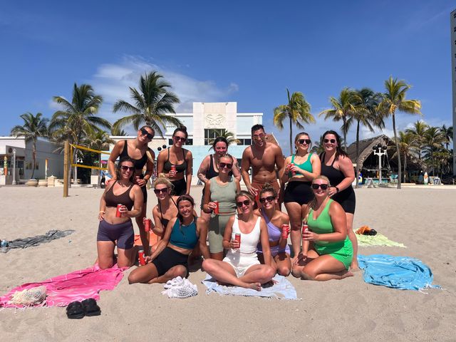 Smiling group of people in colorful swimwear posing with canned drinks on a sunny tropical beach with palm trees, a beach volleyball net and resort buildings under a clear blue sky