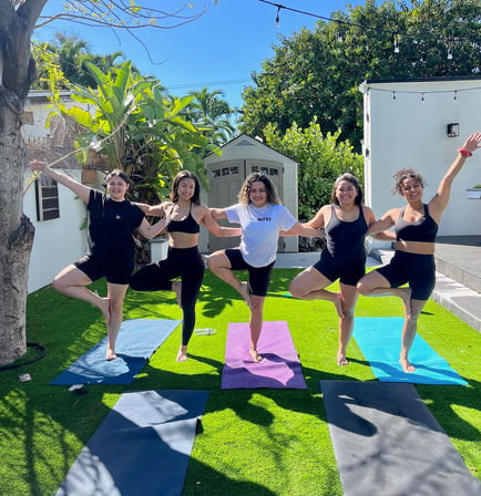 Five women doing tree pose together on colorful yoga mats in a sunny tropical backyard with palm trees and a garden shed, arms linked and smiling.