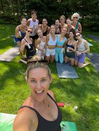 Cheerful group selfie of women in activewear on yoga mats on a sunny green lawn after an outdoor yoga session, smiling and holding canned drinks.