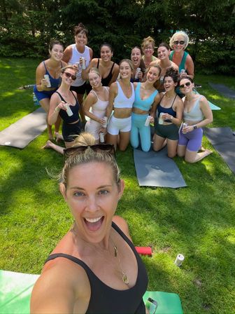 Cheerful group selfie of women in activewear on yoga mats on a sunny green lawn after an outdoor yoga session, smiling and holding canned drinks.