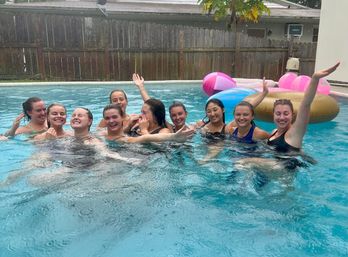 Group of friends laughing and waving in a backyard swimming pool with colorful inflatable floats, rippling water, and a wooden fence and house in the background.