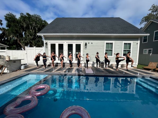 Group of people in black workout clothes doing synchronized yoga on a wooden pool deck in a sunny suburban backyard, light-green house with white trim behind them and inflatable rings floating in the blue swimming pool.