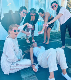 Six women friends on a sunny wooden deck in yoga clothes and pink heart-shaped sunglasses playfully pouring canned drinks over a friend lying on a yoga mat — outdoor girls' day vibe.
