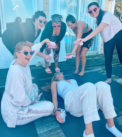 Six women friends on a sunny wooden deck in yoga clothes and pink heart-shaped sunglasses playfully pouring canned drinks over a friend lying on a yoga mat — outdoor girls' day vibe.