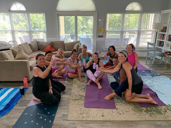 Smiling group of women in workout clothes seated on yoga mats in a bright living-room-style space, toasting with colorful canned drinks after an indoor yoga session