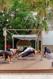 Outdoor yoga class on a wooden deck: group holding plank poses under a pergola with string lights, palm trees, and a rattan swing
