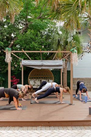 Outdoor yoga class on a wooden deck: group holding plank poses under a pergola with string lights, palm trees, and a rattan swing