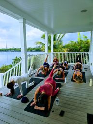 Group yoga class on a covered waterfront deck — students on mats doing leg lifts with palm trees and a calm marina view beyond.