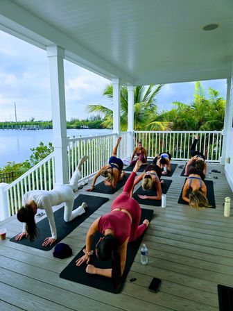 Group yoga class on a covered waterfront deck — students on mats doing leg lifts with palm trees and a calm marina view beyond.