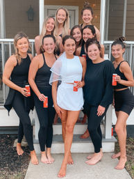 Bachelorette party group photo on a suburban front porch — bride-to-be in white with a sheer veil surrounded by friends in black activewear, all barefoot and smiling on the steps while holding red drink cans.