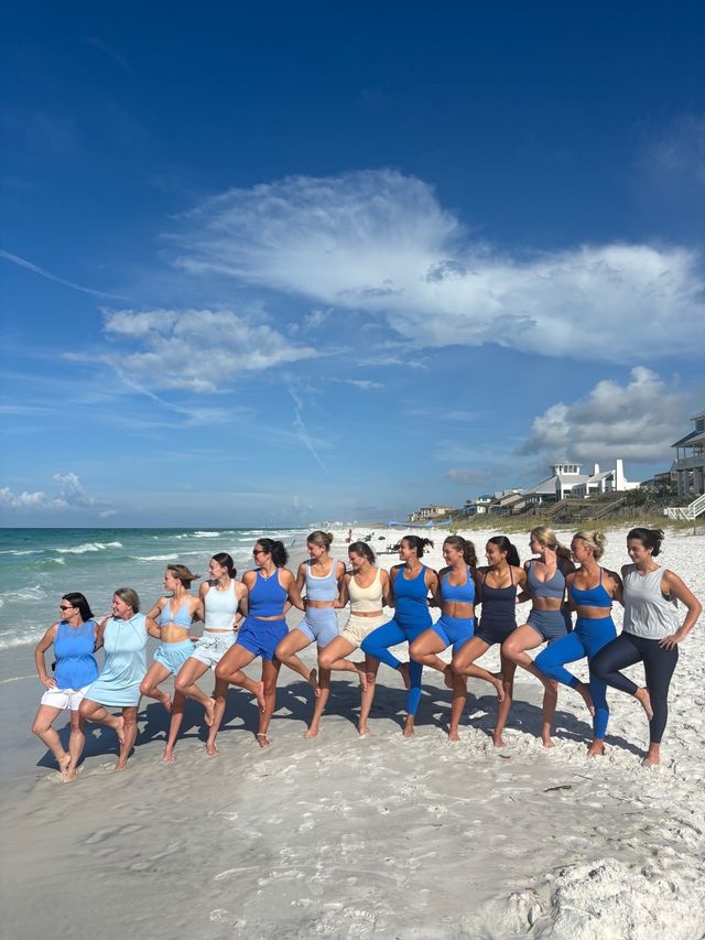 Group of women in blue activewear doing tree-pose on a sunny white-sand beach by turquoise ocean, coastal homes in background and wispy blue sky