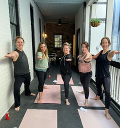Five barefoot women in a group yoga class striking Warrior II on pink mats on a covered urban balcony with exposed brick and a hanging plant.