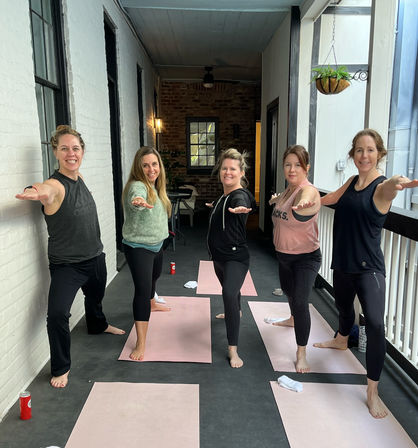Five barefoot women in a group yoga class striking Warrior II on pink mats on a covered urban balcony with exposed brick and a hanging plant.