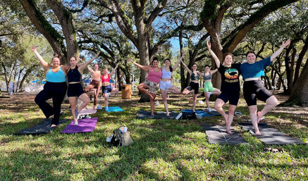 Group of people smiling and holding tree pose on yoga mats during an outdoor class beneath sprawling oak trees in a sunny park