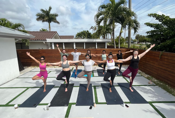 Outdoor group yoga class in a sunny palm-lined backyard, participants on mats holding tree pose