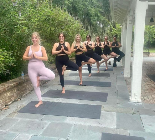 Outdoor yoga class with seven women practicing tree pose on mats along a stone patio under a white pavilion beside lush green foliage.