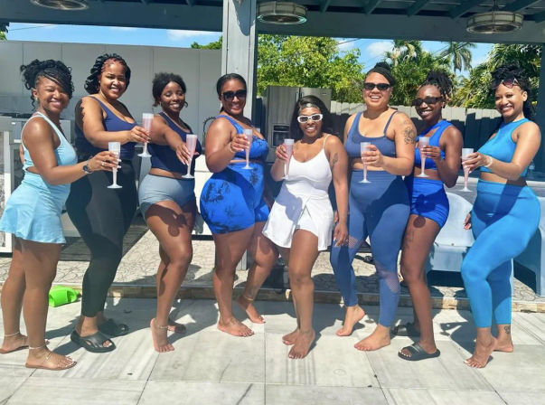 Sun-soaked tropical pool party: eight women in blue and white swimwear smiling and toasting with plastic champagne flutes under a shaded pergola.