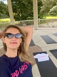 Selfie of a person wearing cat-eye sunglasses and a navy tee with pink script at an outdoor park pavilion set up for yoga — black mats on concrete, a small white cropped tee on one mat, white railing and green trees beyond.