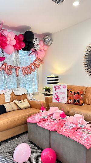 Living room styled for a pink cowgirl girls' party — balloon garland in pink, black and metallic with rose-gold "LAST RODEO" letters, pink cowboy hats, matching pink tees and party cups arranged on leather sofas and ottoman.