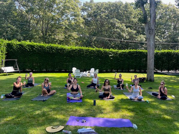 Group of women on yoga mats in a sunny suburban backyard toasting with wine glasses after an outdoor yoga session, with green hedges, trees and Adirondack chairs in the background.