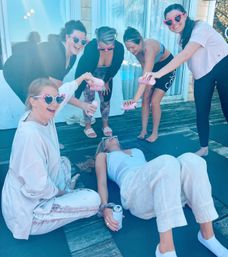 Six friends in heart-shaped sunglasses playfully pour pink canned drinks over a woman lying on yoga mats on a sunny wooden deck by sliding glass doors.